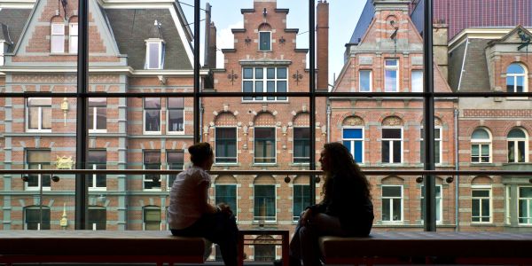 Two people sitting and talking by a large window with a view of old buildings.