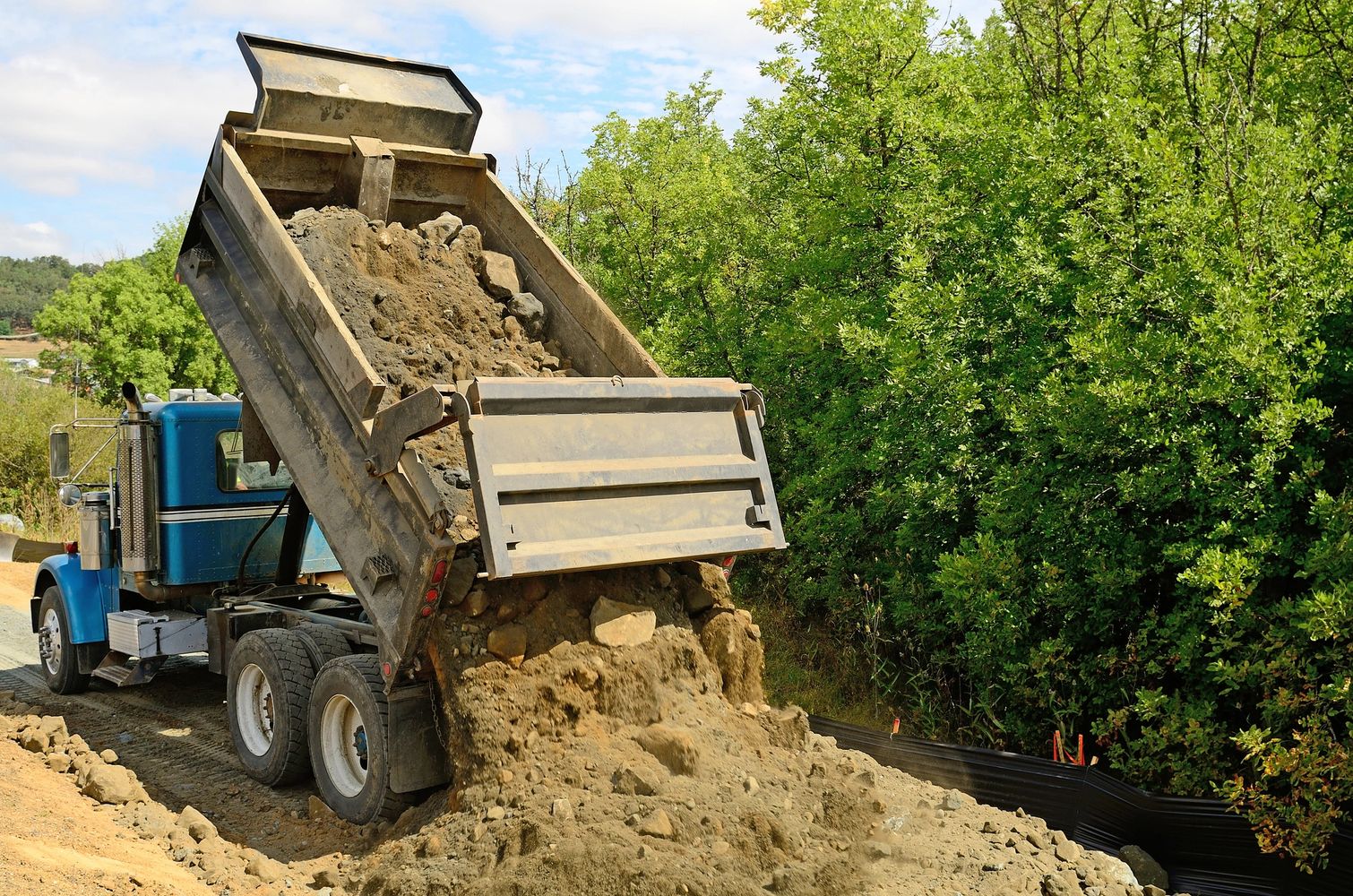 A blue dump truck unloading dirt and rocks at a construction site near green trees.