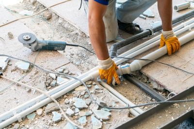 Worker installing PVC pipes on a broken tile floor with tools around.