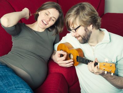 Pregnant woman smiles as man plays ukulele beside her on a red couch.