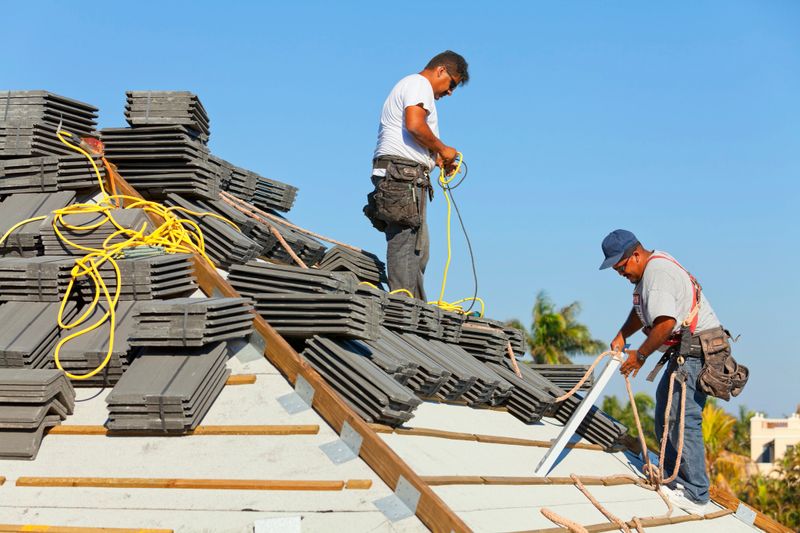 Two roofers preparing to lay roof tile on an estate home. One is plugging in an extension cord for the nail gun, while the other is preparing the eaves riser.