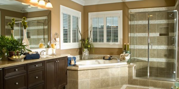 Bright bathroom with glass shower and large bathtub under shuttered windows.