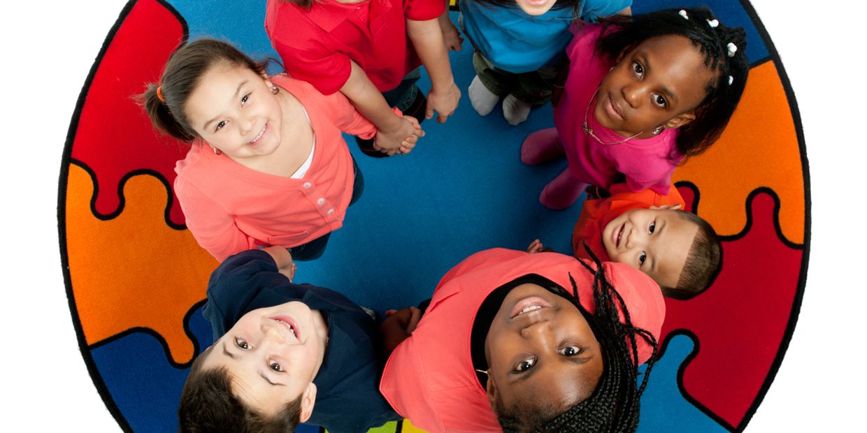 Diverse group of children standing in a circle on a colorful puzzle-piece rug, looking up and smiling.