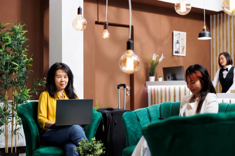 Smiling Asian visitor using laptop at entrance of comfortable resort while on vacation. Traveling guests relaxing in modern hotel lounge area using gadgets during holiday