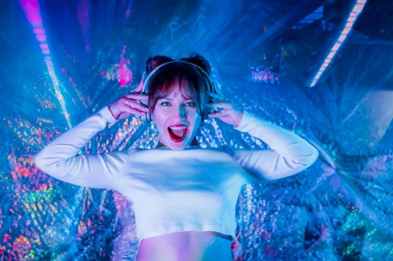 Portrait of emotional screaming woman in white clothes and headphones dancing in neon light. Dj , music lover. White dress code party. Silent disco. Enjoy moment on shiny background. Selective focus