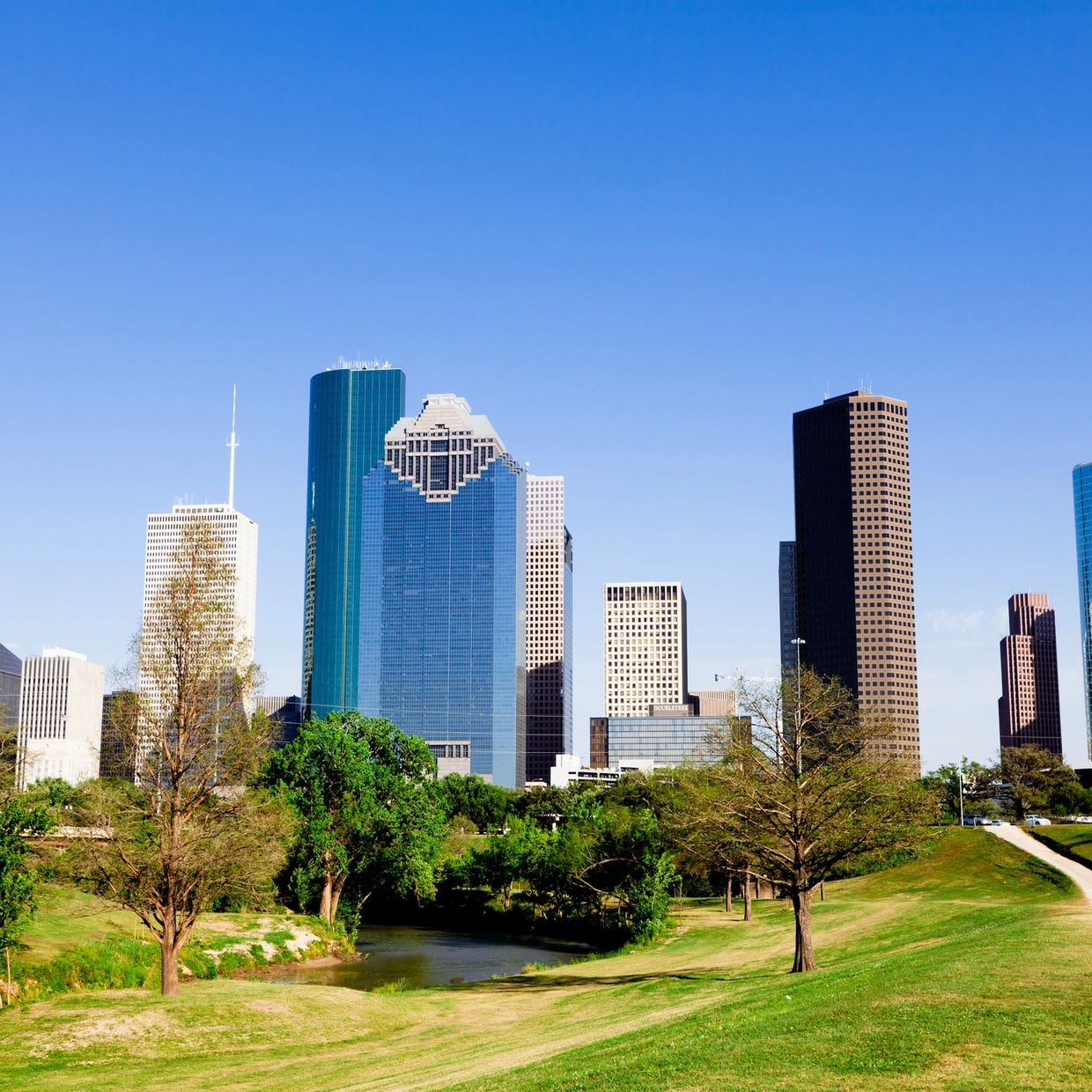 Skyline of Houston with green park and river in foreground under clear blue sky.