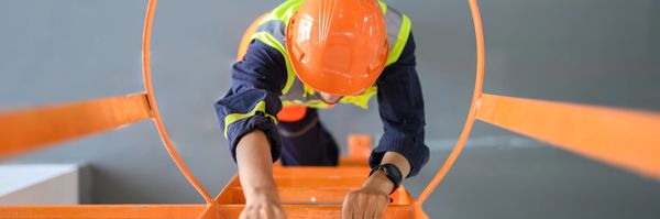 Construction worker climbing an orange safety ladder with a protective cage.