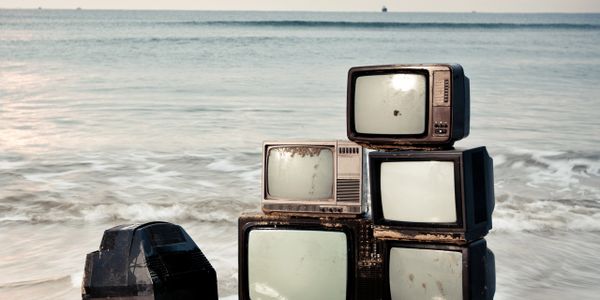 A stack of old, dusty televisions on a beach shore with waves in the background.