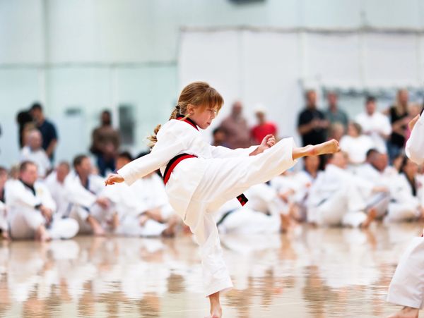 Young girl performs a high kick in a karate class with onlookers in the background.
