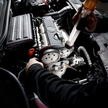Mechanic lubricating a car engine's timing belt area with a grease gun.