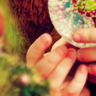 A child's hand holding a colorful ornament near a tree.