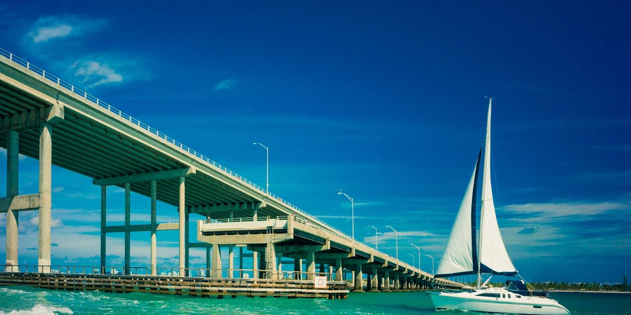 Sailboat gliding beside a long concrete bridge over turquoise water under a clear blue sky.