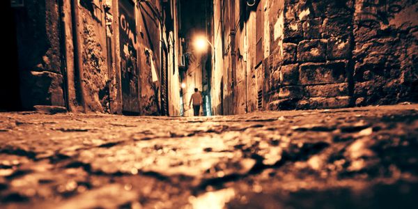 Low-angle view of a person walking down a dimly lit cobblestone alley at night.