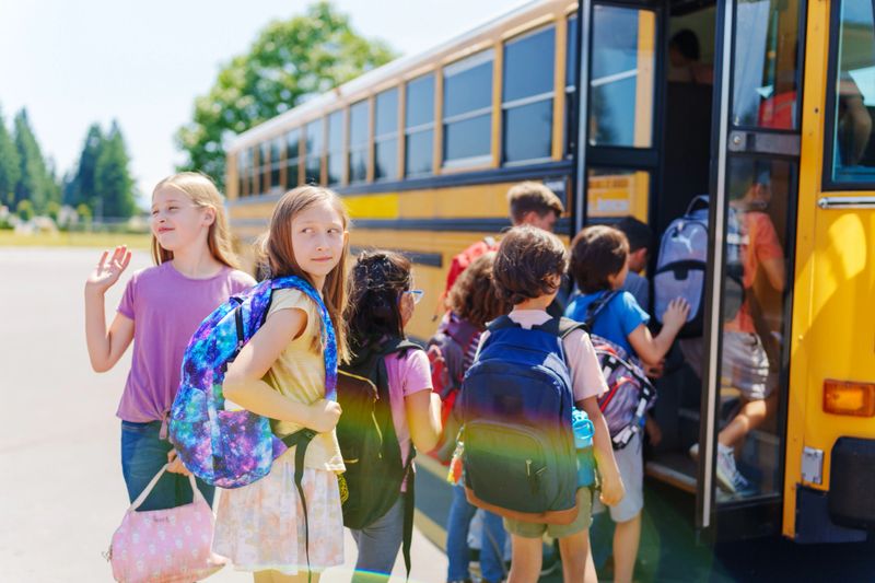 Multiethnic group of elementary age students of varying ages wait in a line while boarding a school bus on a warm and sunny day.