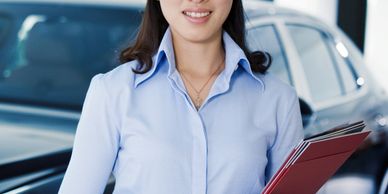 Smiling woman in blue shirt holding documents in front of a car.