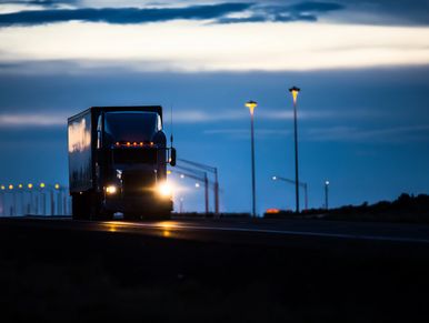 A semi truck drives on a highway at dusk with headlights on.
