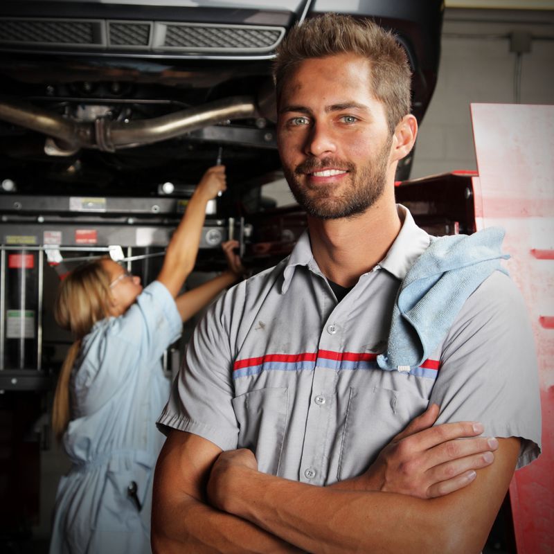 A male mechanic stands and smiles as a female mechanic works on a car.