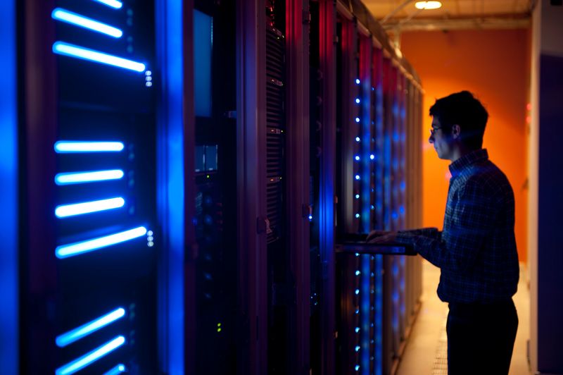The interior of a modern server room in a data center.  An IT engineer is busy configuring the servers. The room is dark, but the servers themselves are lit.  The servers at the left side are lit in blue, while the ones at the other end are lit by an orange light source coming from the right side.