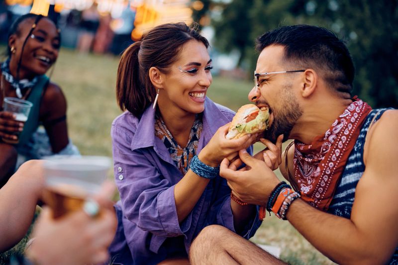 Cheerful woman having fun while feeding her boyfriend with hamburger during open air music concert.