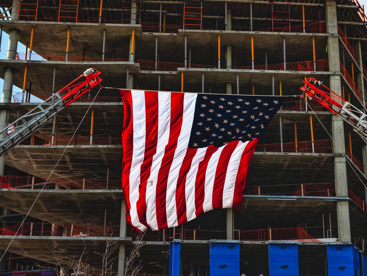 Large American flag hanging between two cranes at a building construction site.