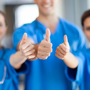 Three healthcare professionals in blue scrubs showing thumbs up.