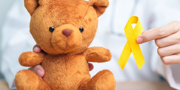 Hand holding yellow ribbon next to a teddy bear, symbolizing childhood cancer awareness.