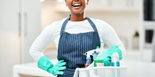Smiling woman in gloves and apron holding cleaning supplies basket.