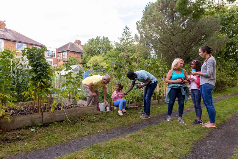 Full shot of a multi-generational family talking and smiling to each other standing next to a family garden full of fresh fruit and vegetables. They are all smiling and talking together wearing casual clothing.