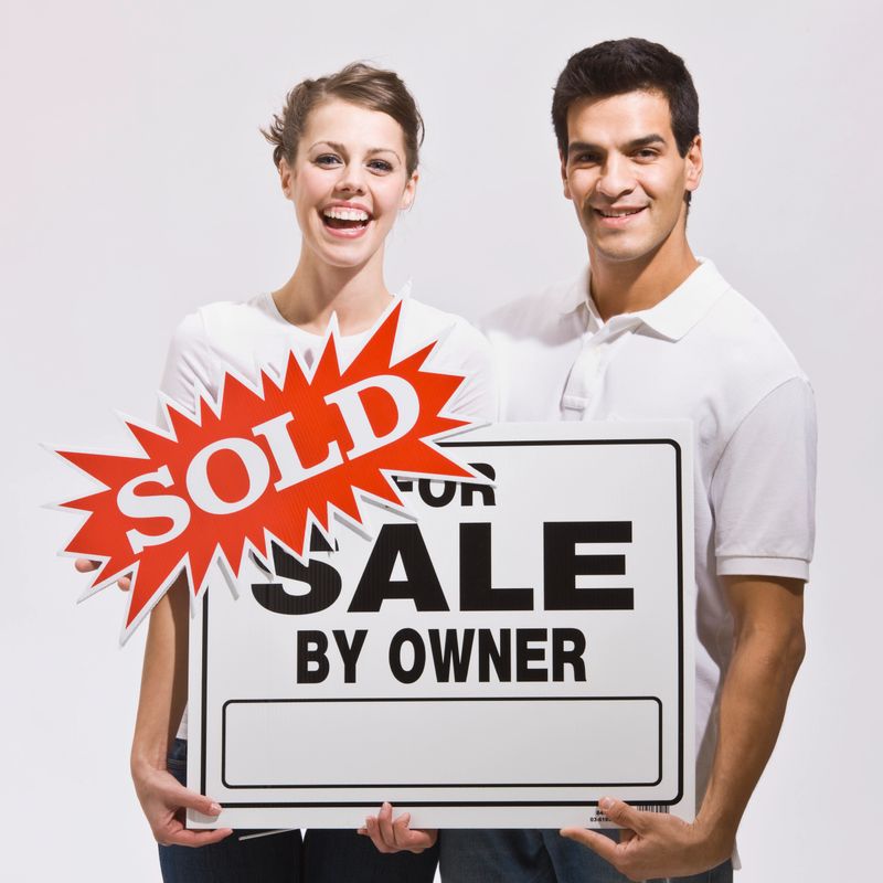 An attractive young couple holding a real estate sign with a 'sold' sticker on it.  They are smiling directly at the camera and look excited. Square.