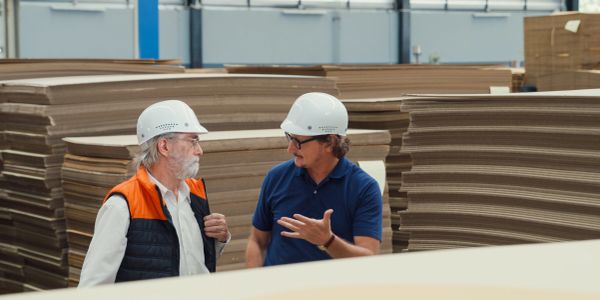 Two men in hard hats discuss in a cardboard factory.