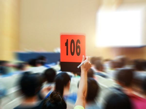 A person holds up a red bidding paddle numbered 106 in a crowded auction room.