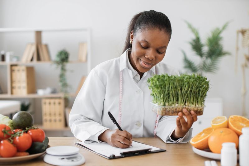 Positive female nutritionist with young sprouts in hand creating meal diet at writing desk in doctor's office. African american specialist with measuring tape recommending clients slimming foods.