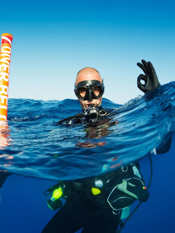 Scuba diver signaling OK next to a safety marker buoy in clear blue water.
