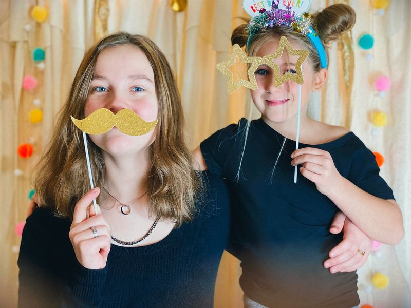 Two girls holding Photo Booth props at a birthday party with a homemade Photo Booth. They are embracing and laughing at the camera.