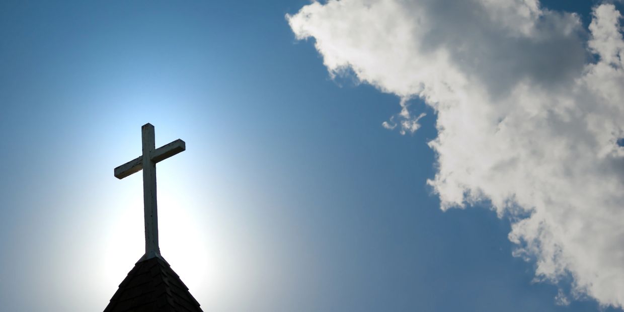 A cross atop a church roof with sun halo and clouds.