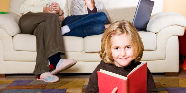 Young girl reading a book on the floor while two women sit on a couch behind her.