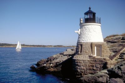 A white lighthouse on rocky shore with a sailboat in the blue sea.