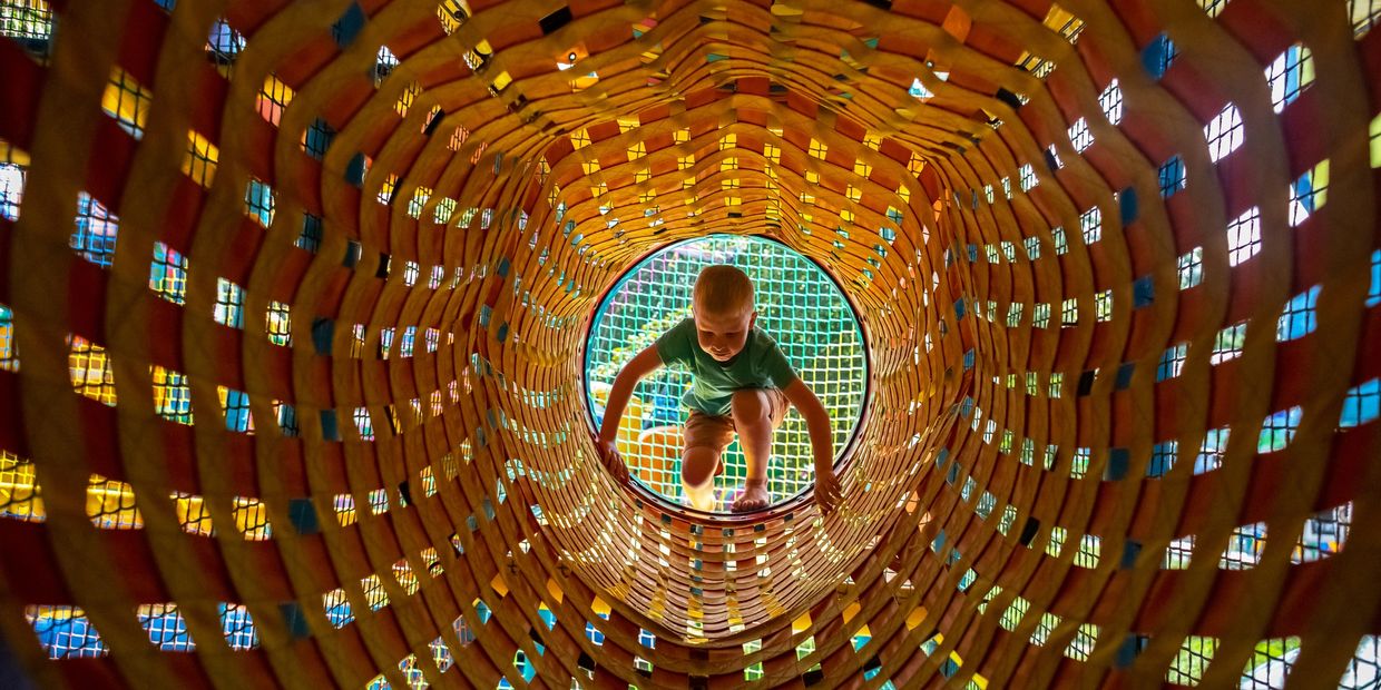 Child climbing through a colorful woven tunnel at a playground.
