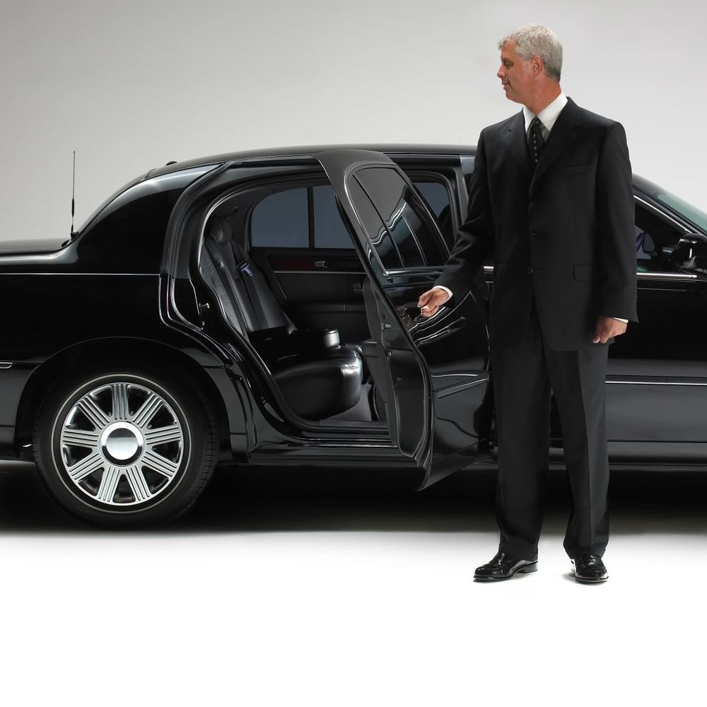 A chauffeur in a black suit opens the door of a luxury black car.