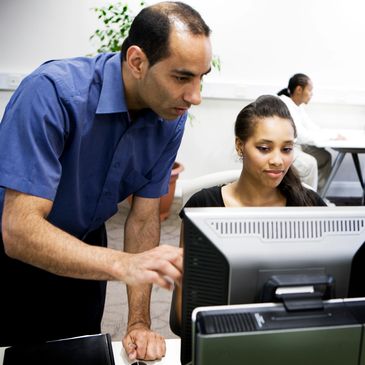 Man in blue shirt helps woman at computer in office setting.