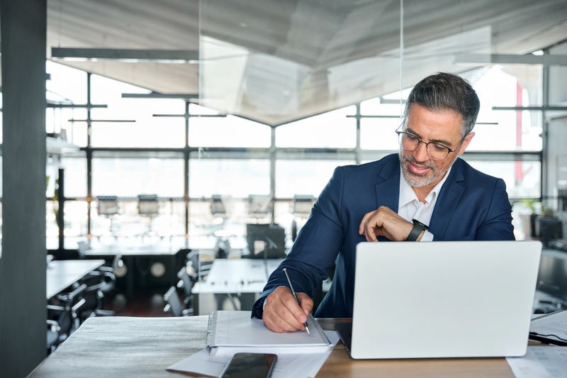 Middle aged smiling professional business man global company executive ceo manager or lawyer wearing suit sitting at desk in modern office working on laptop computer and writing notes, copy space.