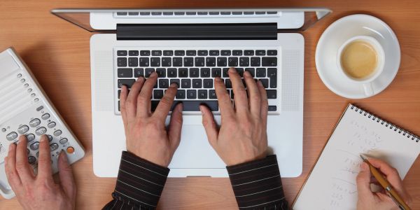 Person multitasking with laptop, calculator, and notepad on wooden desk.