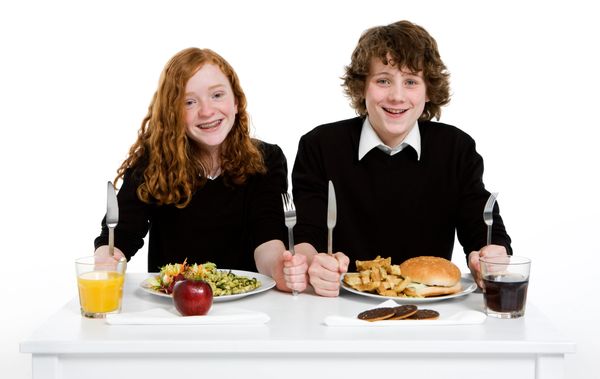 Two kids sitting at a table with different meals, ready to eat.