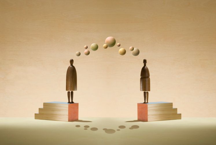 Two wooden figures on blocks exchanging wooden spheres in an arc.