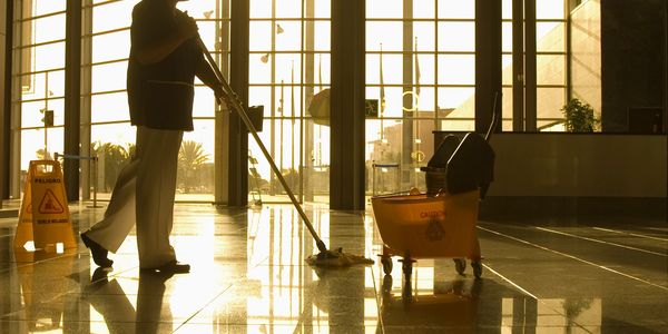 Silhouette of a janitor mopping a shiny floor in a sunlit building lobby.