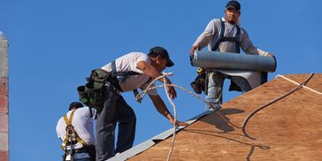 Three construction workers installing roofing material on a sunny day.