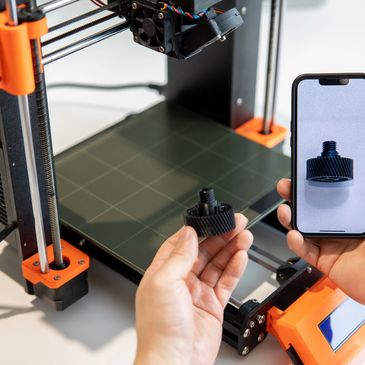 Person holding a 3D printed gear and its photo on a smartphone.
