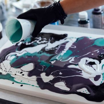 An artist pouring teal paint onto a canvas with abstract patterns.