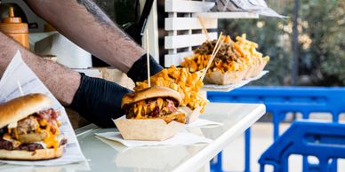 A person serves loaded burgers with fries at an outdoor food stand.