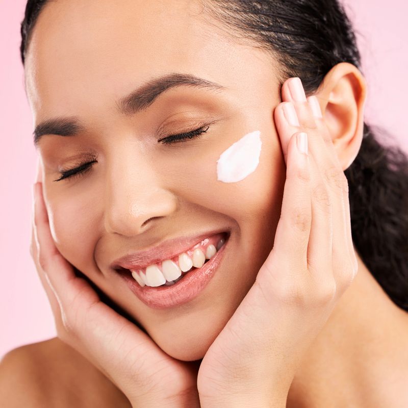 Skincare, happy and woman with face cream in studio isolated on a pink background. Smile, beauty and natural model with moisturizer, cosmetic and apply dermatology product for wellness and health